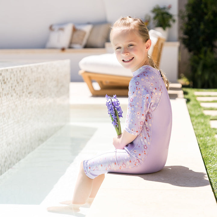 Child wearing oe piece UV sun and sea wetsuit for toddlers in lilac featuring floral print on sleeves, side panels, neck and chest, poolside.
