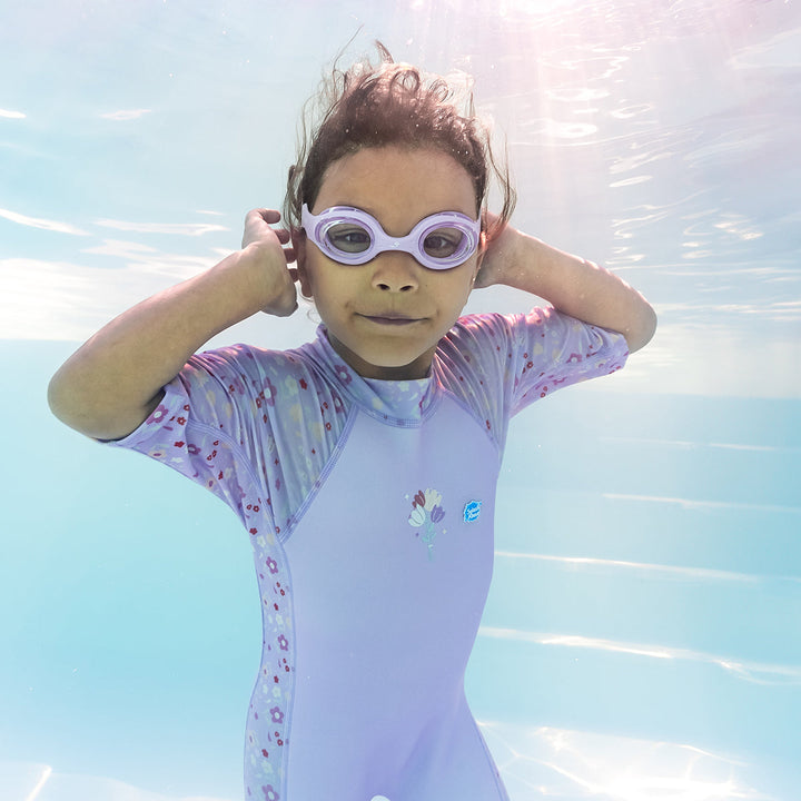 Child wearing oe piece UV sun and sea wetsuit for toddlers in lilac featuring floral print on sleeves, side panels, neck and chest, underwater. Child is also wearing lilac goggles.