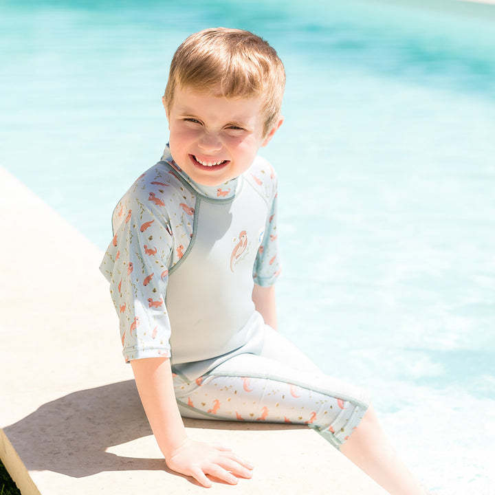 Child wearing one piece UV sun and sea wetsuit for toddlers in light blue, featuring otters themed print on sleeves, side panels, neck and chest. Poolside.