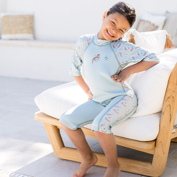 Child wearing one piece UV sun and sea wetsuit for toddlers in light blue, featuring otters themed print on sleeves, side panels, neck and chest.
