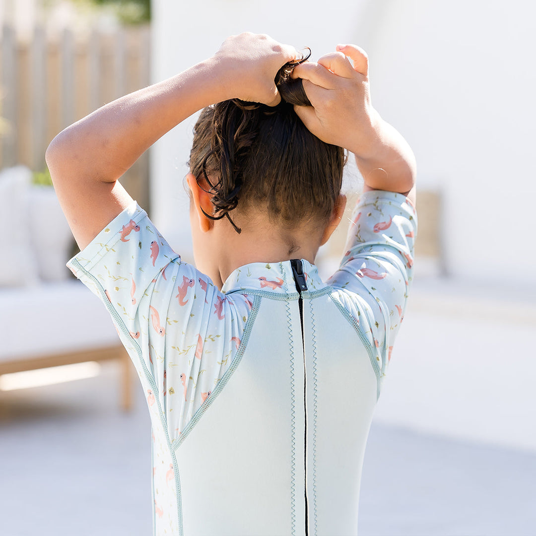 Child wearing one piece UV sun and sea wetsuit for toddlers in light blue, featuring otters themed print on sleeves, side panels, neck and chest. Back