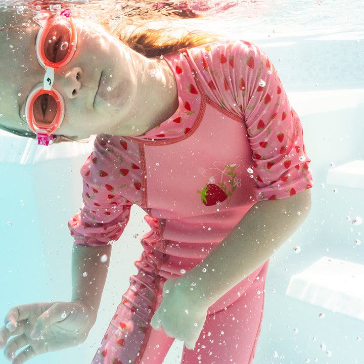 Child swimming in one piece UV sun and sea wetsuit for toddlers in pink, featuring strawberries themed print on sleeves, side panels, neck and chest. Child is also wearing white and pink goggles. Underwater.