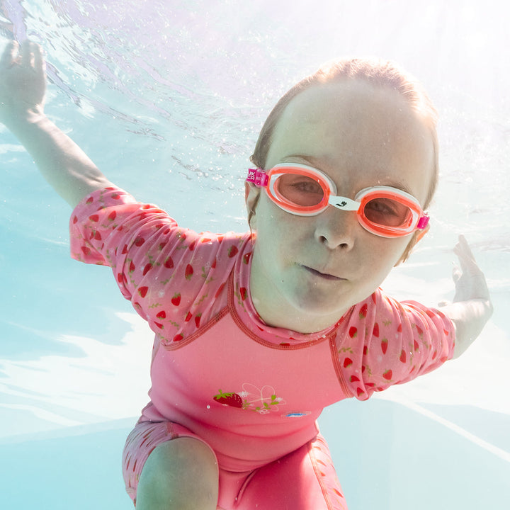 Child swimming in one piece UV sun and sea wetsuit for toddlers in pink, featuring strawberries themed print on sleeves, side panels, neck and chest. Child is also wearing white and pink goggles. Underwater.