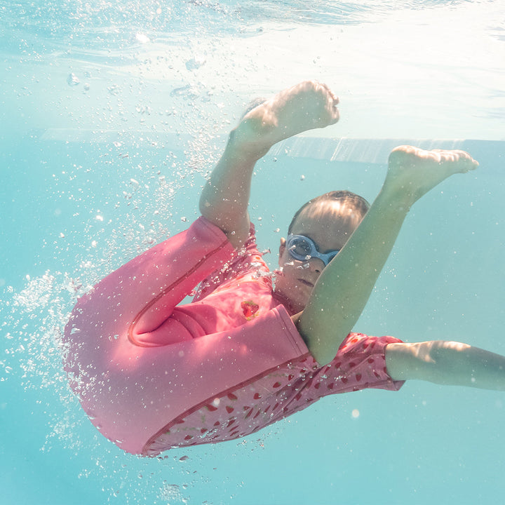 Child swimming in one piece UV sun and sea wetsuit for toddlers in pink, featuring strawberries themed print on sleeves, side panels, neck and chest. Child is also wearing white and pink goggles. Underwater.