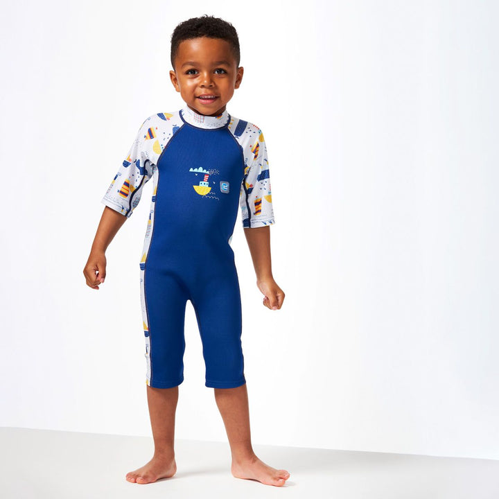 Lifestyle image of child wearing a one piece UV sun and sea wetsuit for toddlers in white and navy. Boats themed print on sleeves, side panels, neck and chest. Front.