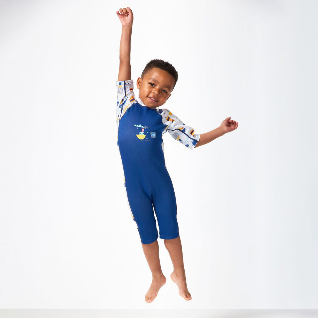 Lifestyle image of child jumping while wearing a one piece UV sun and sea wetsuit for toddlers in white and navy. Boats themed print on sleeves, side panels, neck and chest. Front.