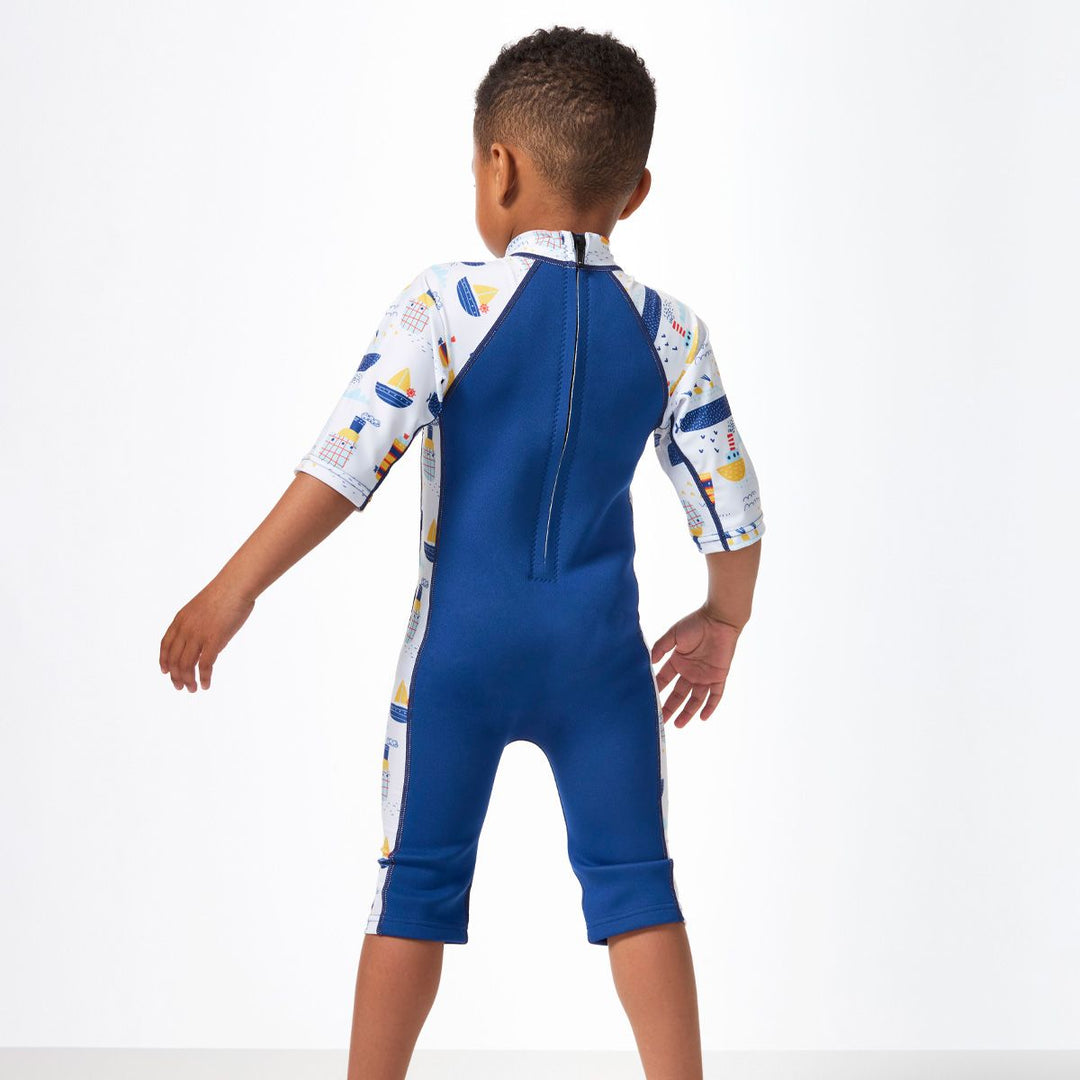 Lifestyle image of child wearing a one piece UV sun and sea wetsuit for toddlers in white and navy. Boats themed print on sleeves, side panels, neck and chest. Back.