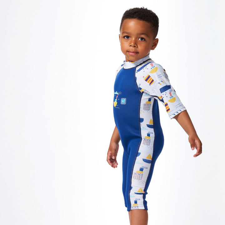 Lifestyle image of child wearing a one piece UV sun and sea wetsuit for toddlers in white and navy. Boats themed print on sleeves, side panels, neck and chest. Side.