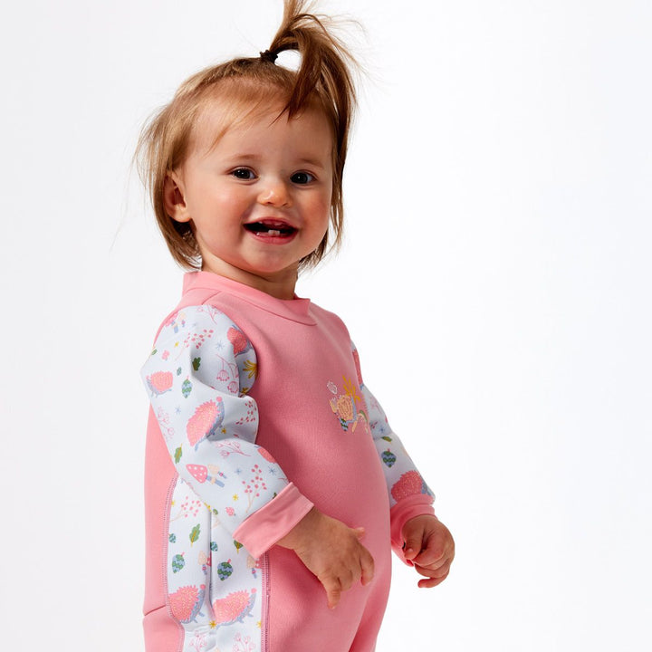 Little one wearing a fleece-lined baby wetsuit in pink and white with pink trims and forest themed print, including hedgehogs, mushrooms, flowers and leaves. Side.