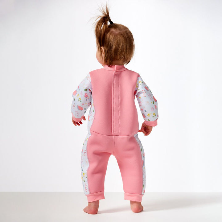 Little one wearing a fleece-lined baby wetsuit in pink and white with pink trims and forest themed print, including hedgehogs, mushrooms, flowers and leaves. Back.