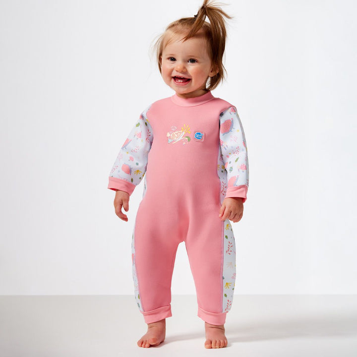 Little one wearing a fleece-lined baby wetsuit in pink and white with pink trims and forest themed print, including hedgehogs, mushrooms, flowers and leaves. Back.