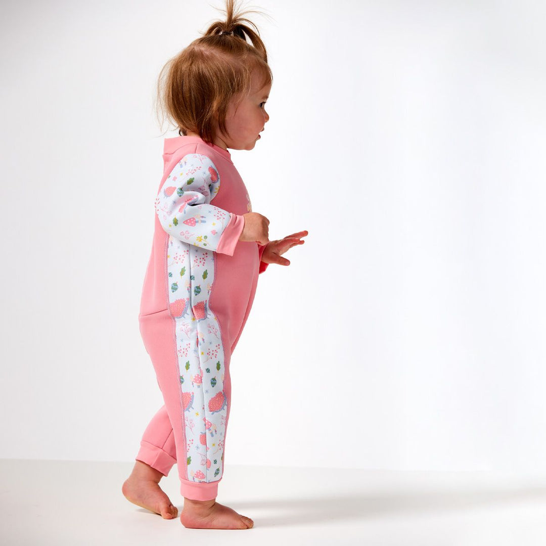 Little one wearing a fleece-lined baby wetsuit in pink and white with pink trims and forest themed print, including hedgehogs, mushrooms, flowers and leaves. Side.