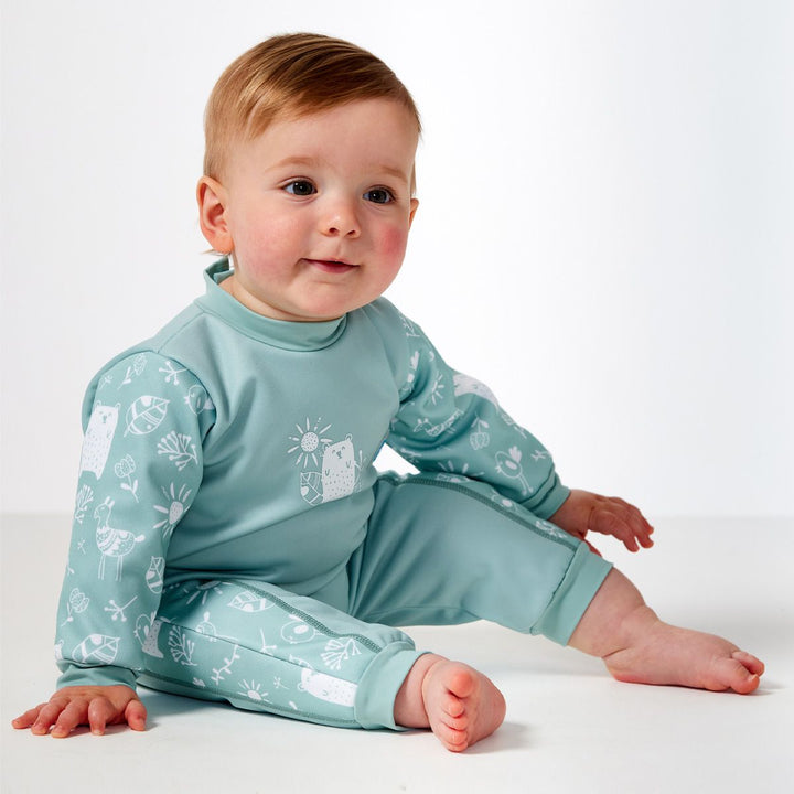 Little one wearing fleece-lined baby wetsuit in greenish blue and white with animals themed print, including bears, llamas, suns and flowers on arms, side panels and chest. Side.