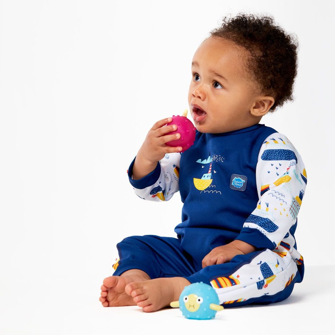 Little one wearing fleece-lined baby wetsuit in navy and white with boats themed print on arms, side panels and chest. Front.