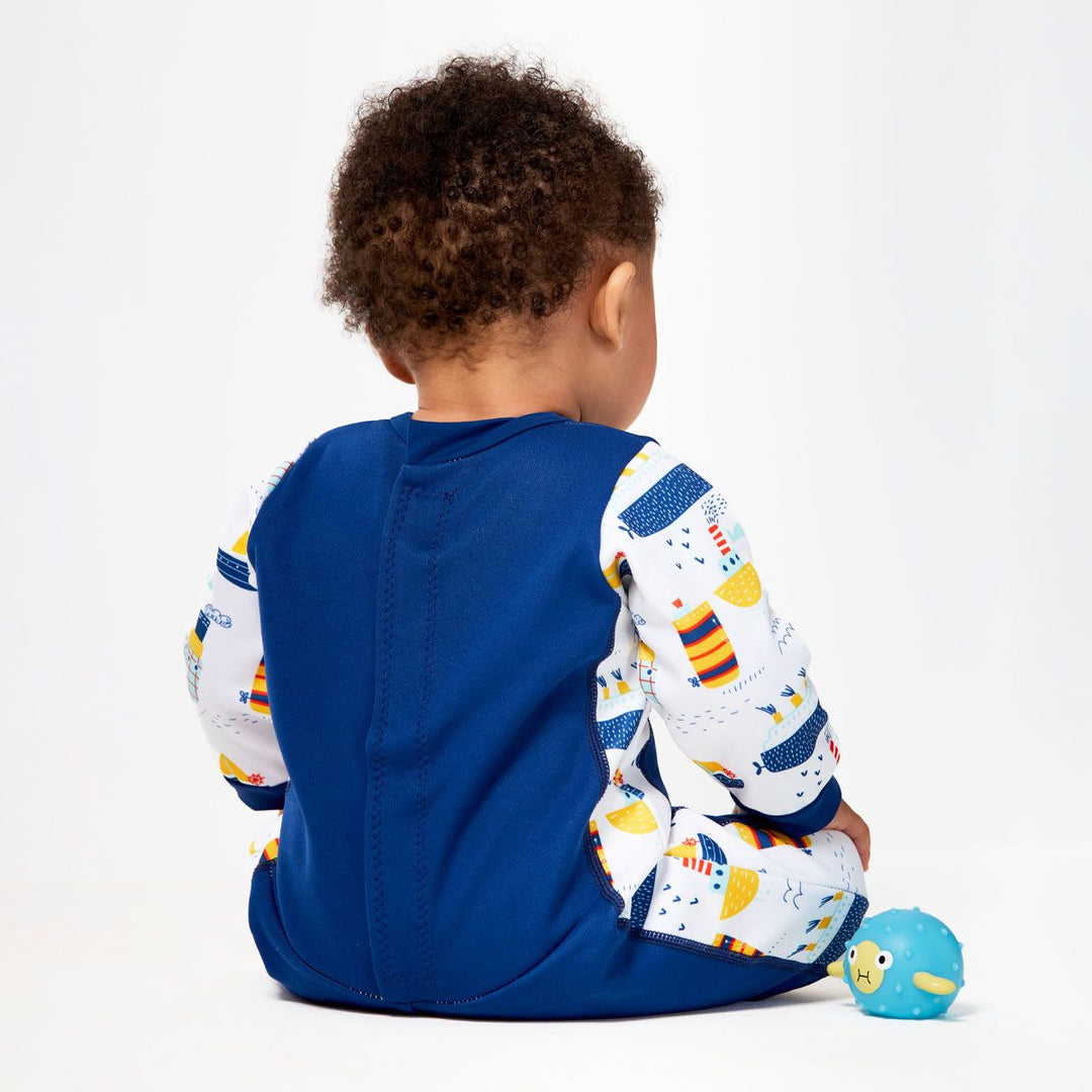 Little one wearing fleece-lined baby wetsuit in navy and white with boats themed print on arms, side panels and chest. Back.