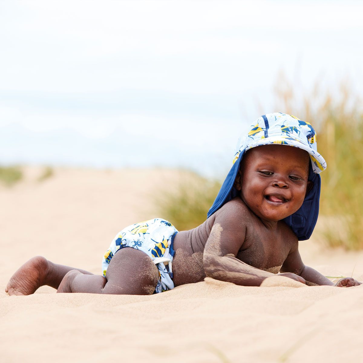 Adjustable under nappy in light blue with navy blue trims and cute bugs print. Lifestyle image of baby in the beach.