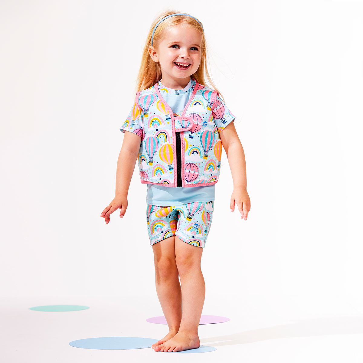 Lifestyle image of child wearing neoprene swim vest for toddlers with non-removable floats in baby blue, pink trims and hot air balloons themed print, including clouds and rainbows. She's also wearing matching jammers and rash top.