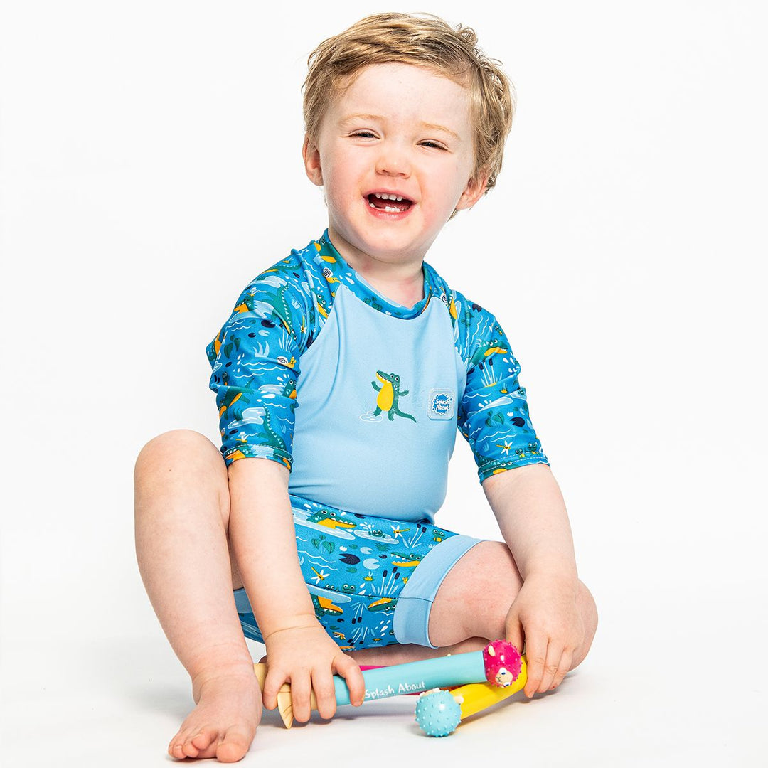 Lifestyle image of toddler wearing a Happy Nappy Sunsuit in blue and swamp themed print, including crocodiles, snails, fireflies, frogs and more. Front.