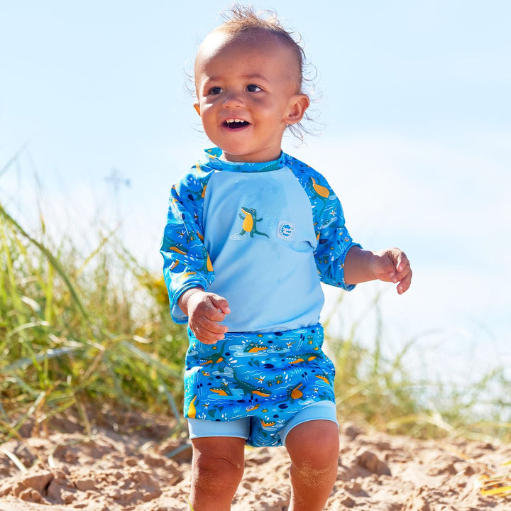 Lifestyle image of toddler wearing a Happy Nappy Sunsuit in blue and swamp themed print, including crocodiles, snails, fireflies, frogs and more. Front.