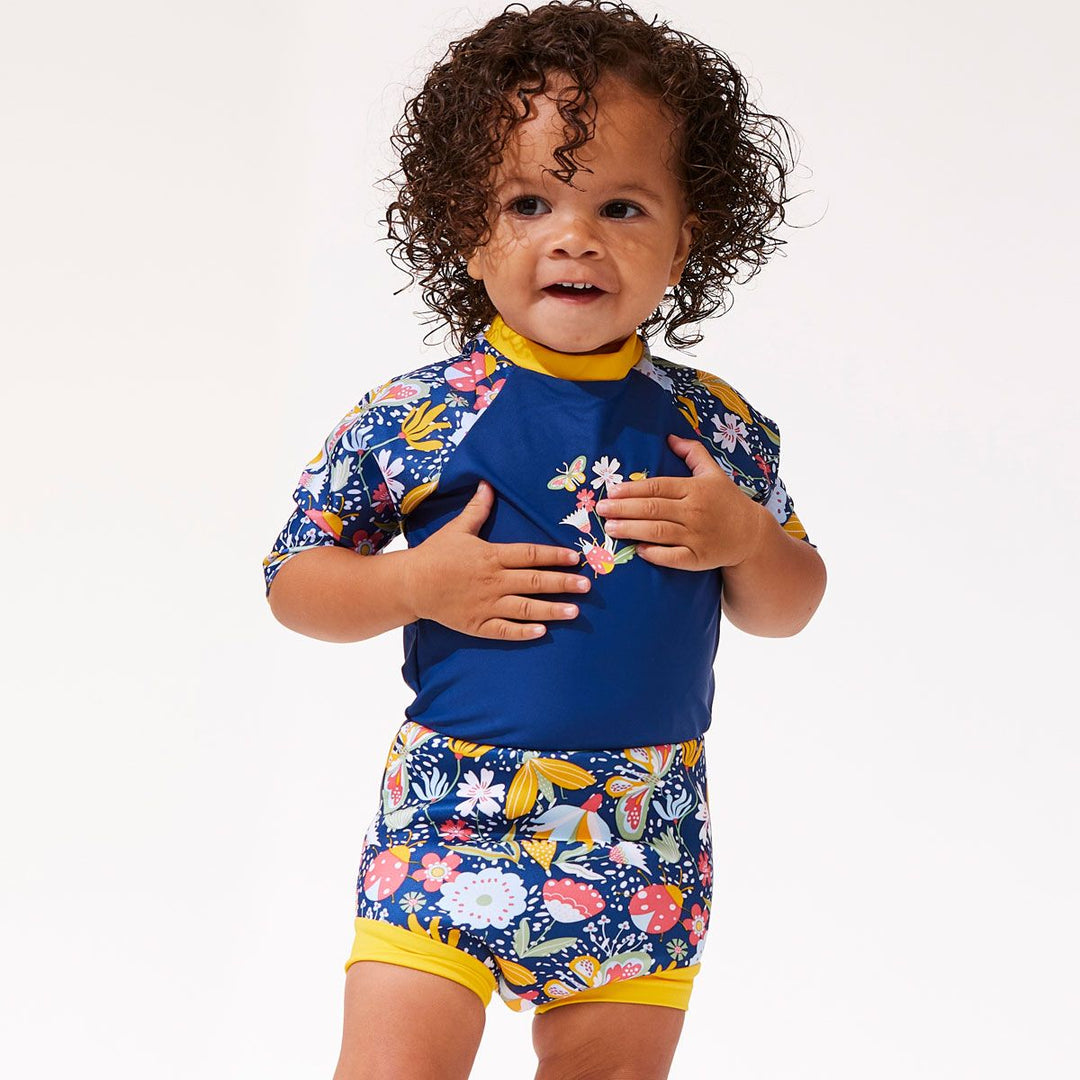 Lifestyle image of toddler wearing a Happy Nappy Sunsuit in navy blue with yellow trims and floral print. Front.