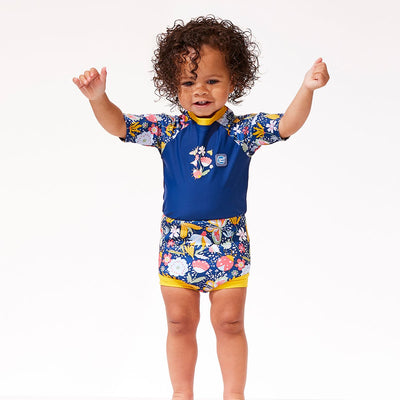 Lifestyle image of toddler wearing a Happy Nappy Sunsuit in navy blue with yellow trims and floral print. Front.