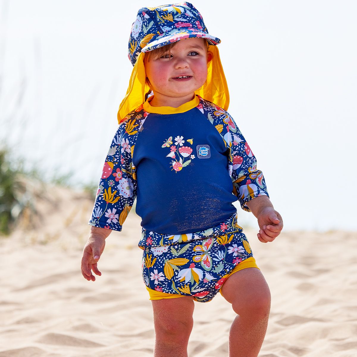 Lifestyle image of toddler wearing a Happy Nappy Sunsuit in navy blue with yellow trims and floral print. Front. He's also wearing a matching legionnaire hat.