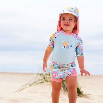 Lifestyle image of child wearing a Happy Nappy Sunsuit in baby blue with pink trims and hot air balloons themed print, including rainbows and clouds. Front. She's also wearing a matching legionnaire hat.