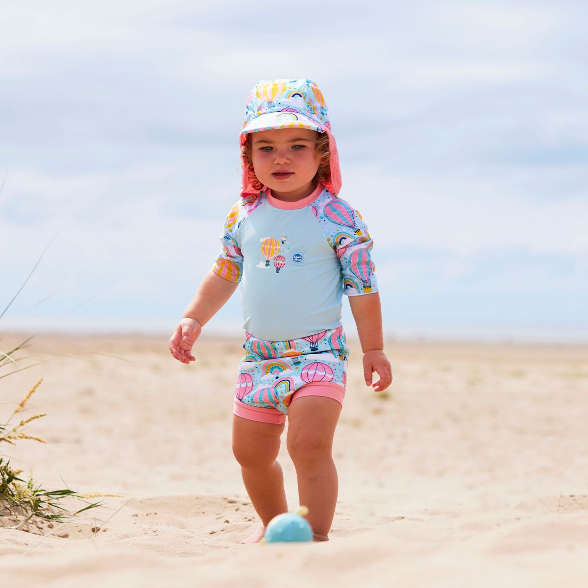 Lifestyle image of child wearing a Happy Nappy Sunsuit in baby blue with pink trims and hot air balloons themed print, including rainbows and clouds. Front. She's also wearing a matching legionnaire hat.