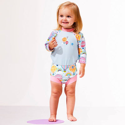 Lifestyle image of child wearing a Happy Nappy Sunsuit in baby blue with pink trims and hot air balloons themed print, including rainbows and clouds. Front.