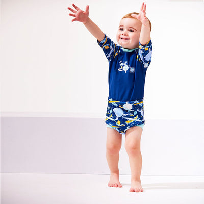 Lifestyle image of toddler wearing a Happy Nappy Sunsuit in navy blue and sky themed print, including airplanes, kites, clouds and hot air balloons. Front.
