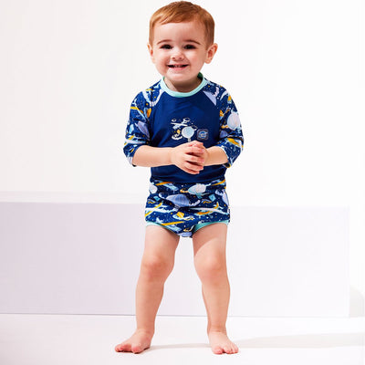 Lifestyle image of toddler wearing a Happy Nappy Sunsuit in navy blue and sky themed print, including airplanes, kites, clouds and hot air balloons. Front.