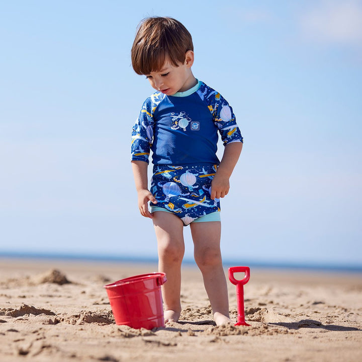 Lifestyle image of child wearing a Happy Nappy Sunsuit in navy blue and sky themed print, including airplanes, kites, clouds and hot air balloons. Front.