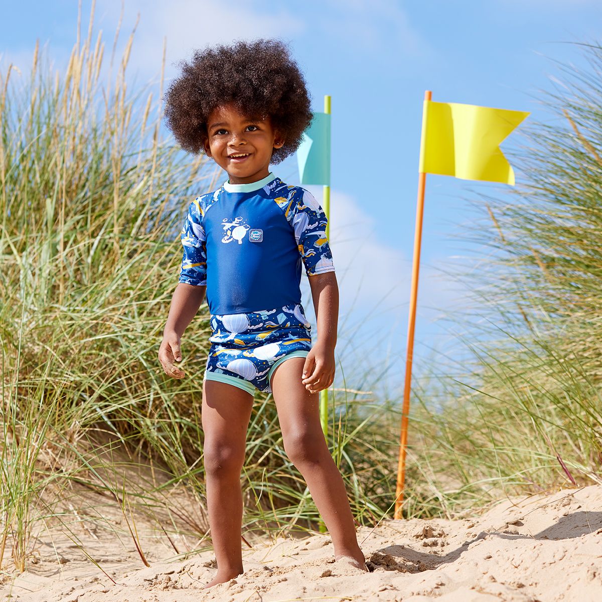 Lifestyle image of child wearing a Happy Nappy Sunsuit in navy blue and sky themed print, including airplanes, kites, clouds and hot air balloons. Front.