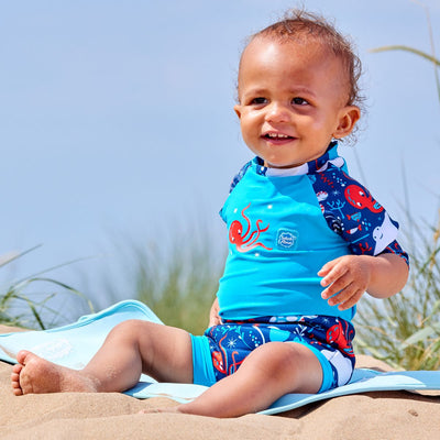 Lifestyle image of toddler wearing a Happy Nappy Sunsuit in navy blue and cyan, with under the sea themed print, including turtles, stingrays, octopus, fish and more. Front.