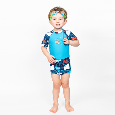 Lifestyle image of toddler wearing a Happy Nappy Sunsuit in navy blue and cyan, with under the sea themed print, including turtles, stingrays, octopus, fish and more. Front. He's also wearing Guppy goggles in blue and green.