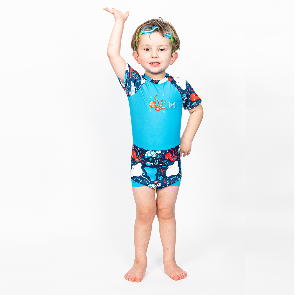 Lifestyle image of toddler wearing a Happy Nappy Sunsuit in navy blue and cyan, with under the sea themed print, including turtles, stingrays, octopus, fish and more. Front. He's also wearing Guppy goggles in blue and green.