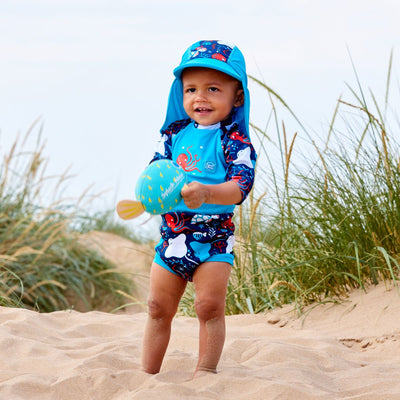 Lifestyle image of toddler wearing a Happy Nappy Sunsuit in navy blue and cyan, with under the sea themed print, including turtles, stingrays, octopus, fish and more. Front. He's also wearing matching legionnaire hat.