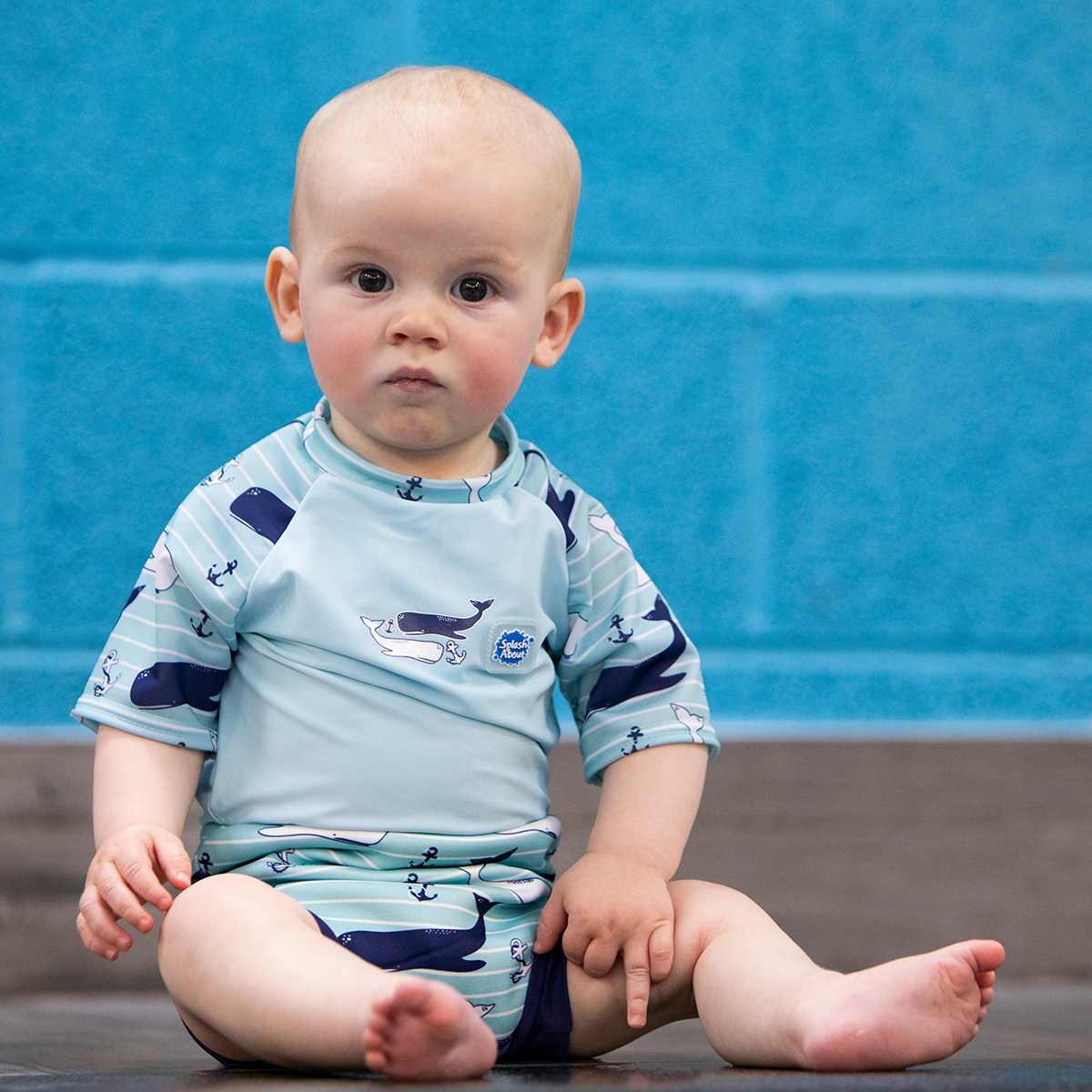 Lifestyle image of baby wearing a Happy Nappy Sunsuit in light blue and whales themed print. Front.