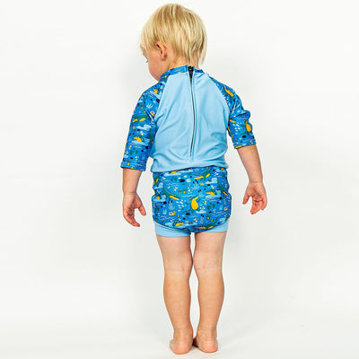 Lifestyle image of toddler wearing a Happy Nappy Sunsuit in blue and swamp themed print, including crocodiles, snails, fireflies, frogs and more. Back.