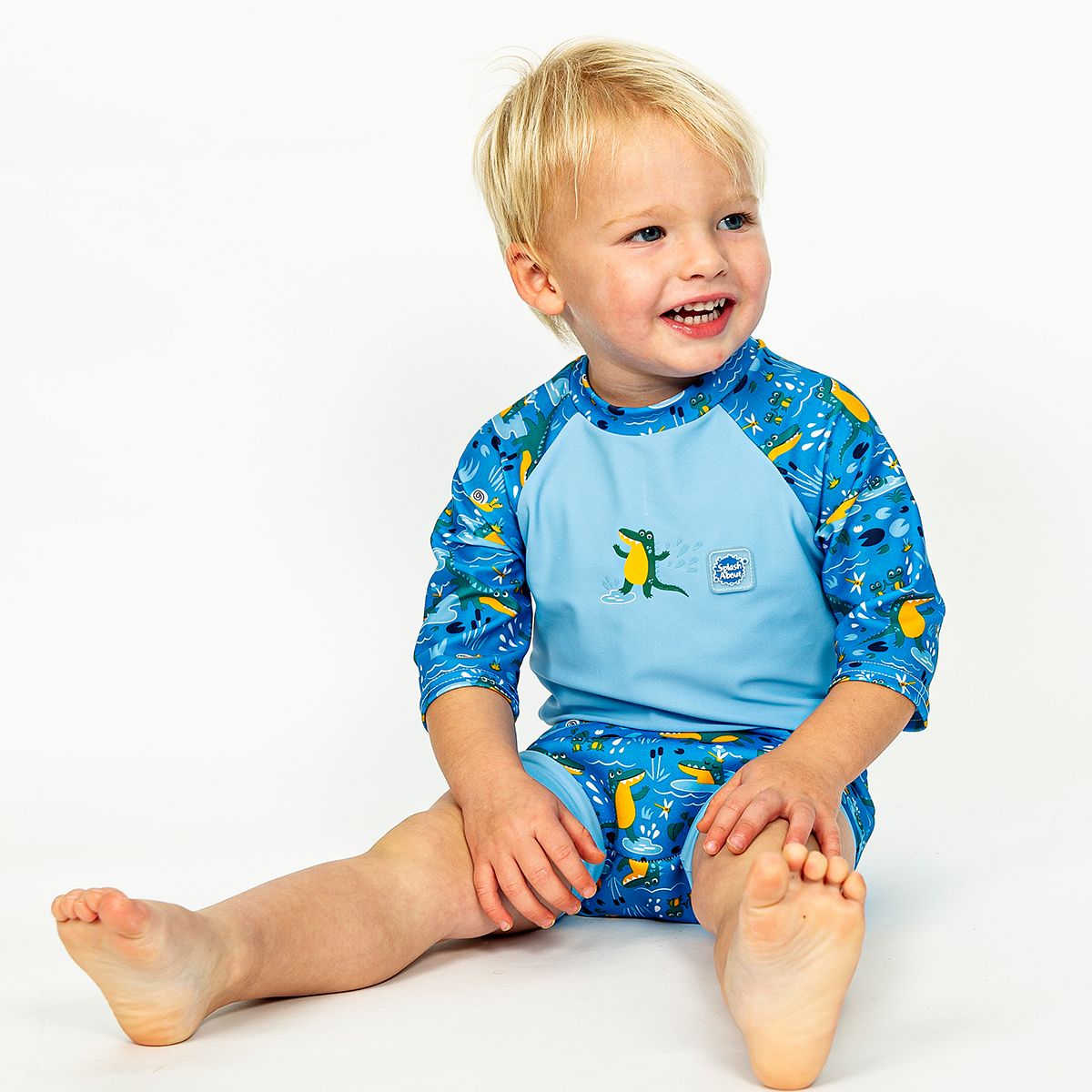 Lifestyle image of toddler wearing a Happy Nappy Sunsuit in blue and swamp themed print, including crocodiles, snails, fireflies, frogs and more. Front.