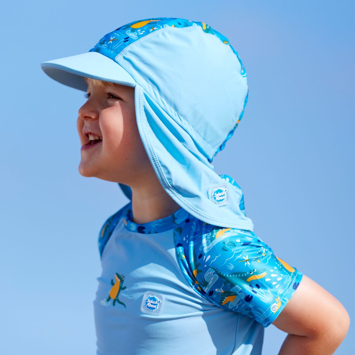 Lifestyle image of toddler wearing a legionnaire style sun hat in the beach. The hat is light blue and blue, with swamp themed print panel including crocodiles, frogs, fireflies and more. He's also wearing matching rash top.
