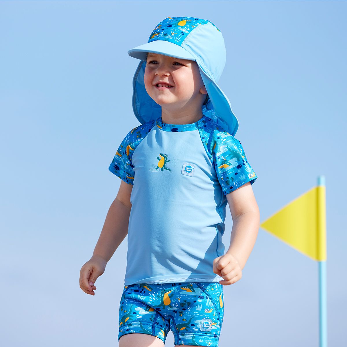 Lifestyle image of toddler wearing a legionnaire style sun hat in the beach. The hat is light blue and blue, with swamp themed print panel including crocodiles, frogs, fireflies and more. He's also wearing matching rash top and jammers.