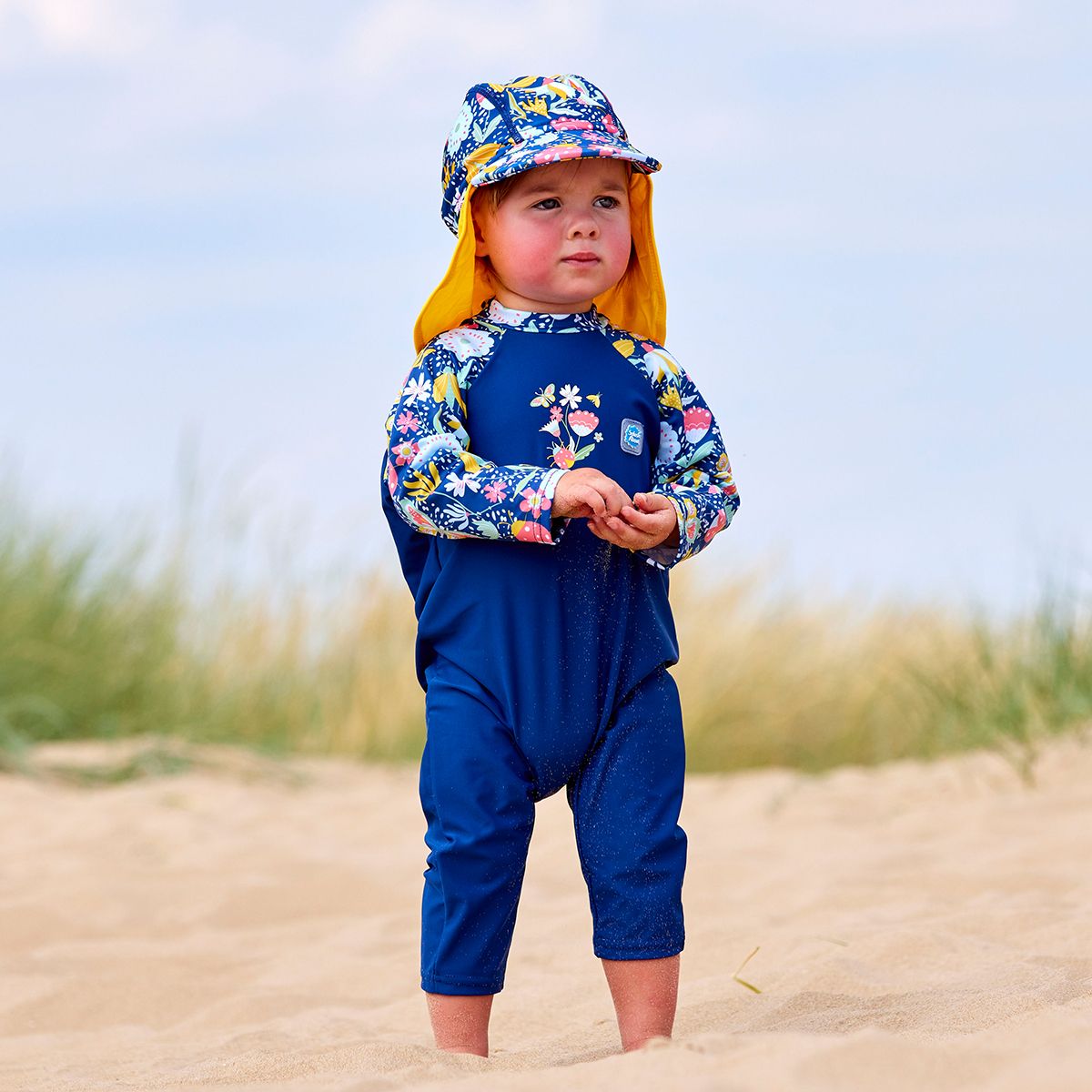 Lifestyle image of toddler wearing a legionnaire style sun hat in the beach. The hat is navy blue, with yellow trims and floral print panel . He's also wearing matching UV All in One sunsuit. Front.