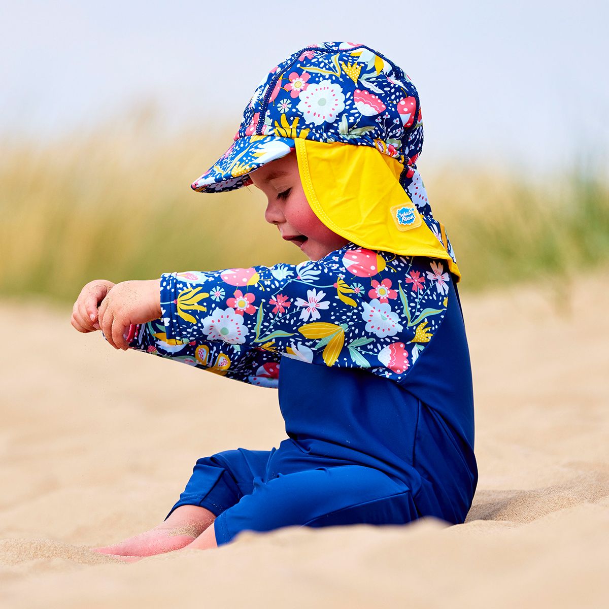 Lifestyle image of toddler wearing a legionnaire style sun hat in the beach. The hat is navy blue, with yellow trims and floral print panel . He's also wearing matching UV All in One sunsuit.
