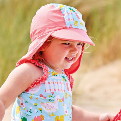 Lifestyle image of toddler wearing a legionnaire style sun hat in the beach. The hat is reddish pink and light blue, with little ducks themed print panel including ducks, snails, flowers and more. She's also wearing matching Happy Nappy costume. Close-up.