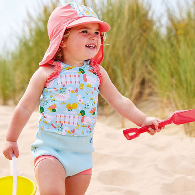 Lifestyle image of toddler wearing a legionnaire style sun hat in the beach. The hat is reddish pink and light blue, with little ducks themed print panel including ducks, snails, flowers and more. She's also wearing matching Happy Nappy costume. Front.