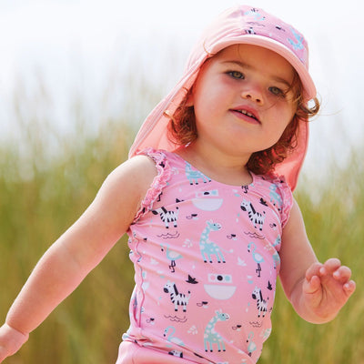 Lifestyle image of toddler wearing a legionnaire style sun hat in the beach. The hat is baby pink, with Noah's Ark themed print panel including giraffes, zebras, flamingos and more. She's also wearing matching Happy Nappy costume. Front.