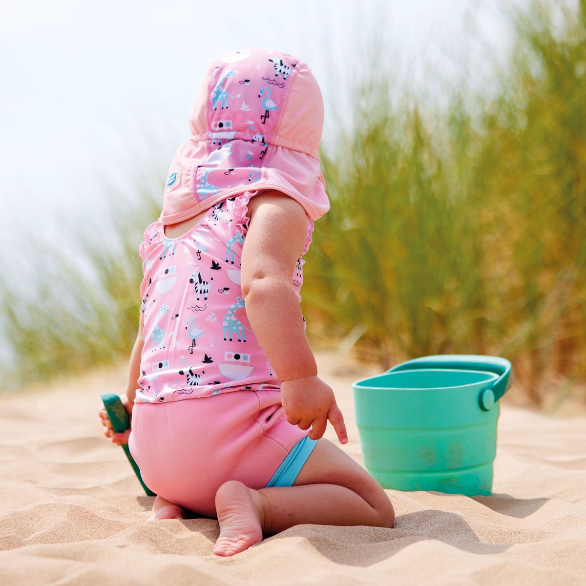 Lifestyle image of toddler wearing a legionnaire style sun hat in the beach. The hat is baby pink, with Noah's Ark themed print panel including giraffes, zebras, flamingos and more. She's also wearing matching Happy Nappy costume. Back.