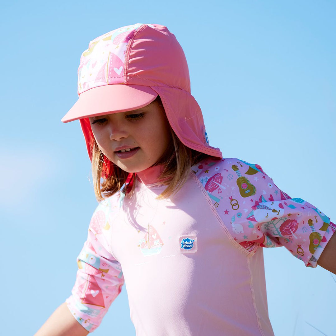 Lifestyle image of toddler wearing a legionnaire style sun hat in the beach. The hat is reddish pink and baby pink, with the owl and the pussycat themed print panel. She's also wearing matching sun and sea wetsuit. Close-up.