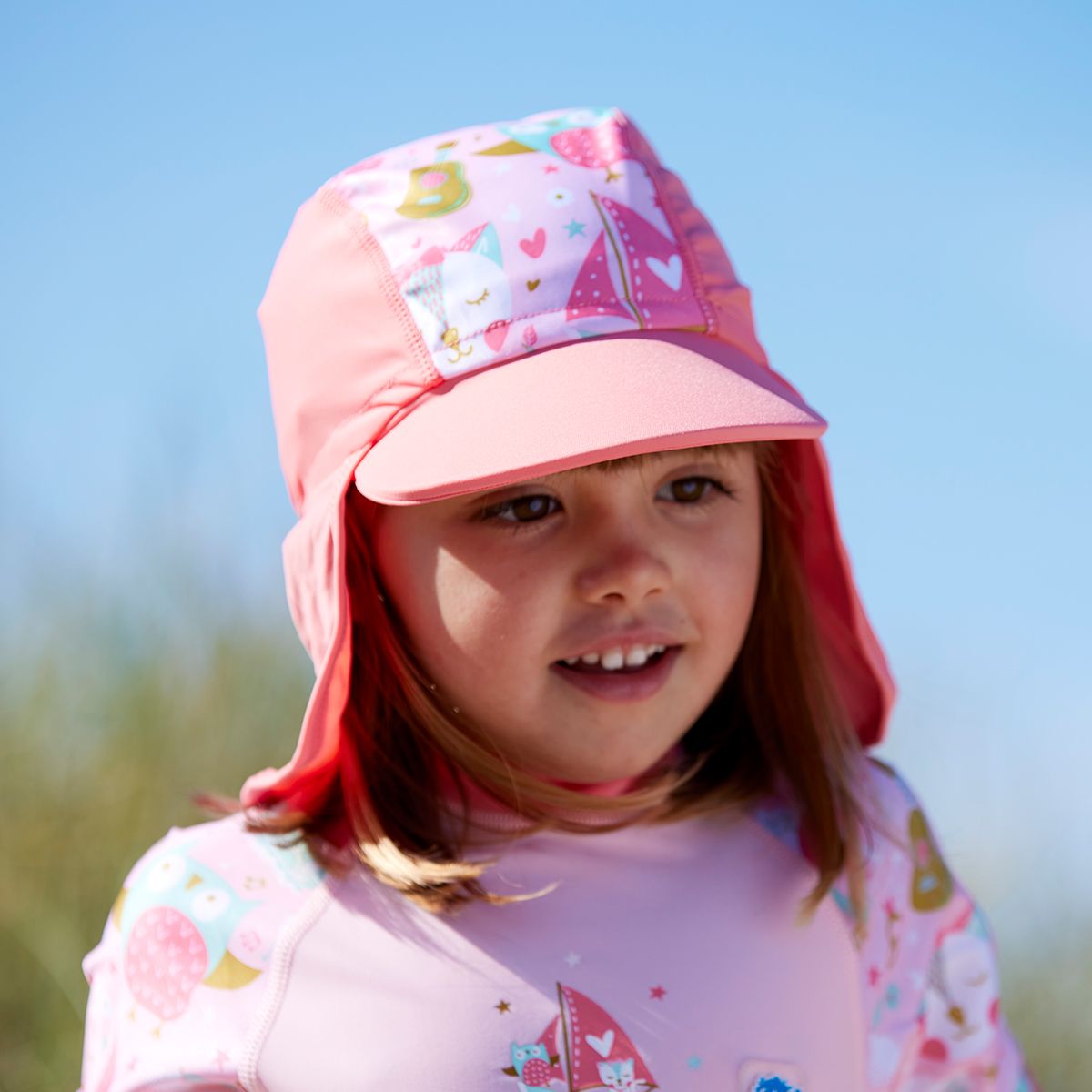 Lifestyle image of toddler wearing a legionnaire style sun hat in the beach. The hat is reddish pink and baby pink, with the owl and the pussycat themed print panel. She's also wearing matching sun and sea wetsuit. Close-up.